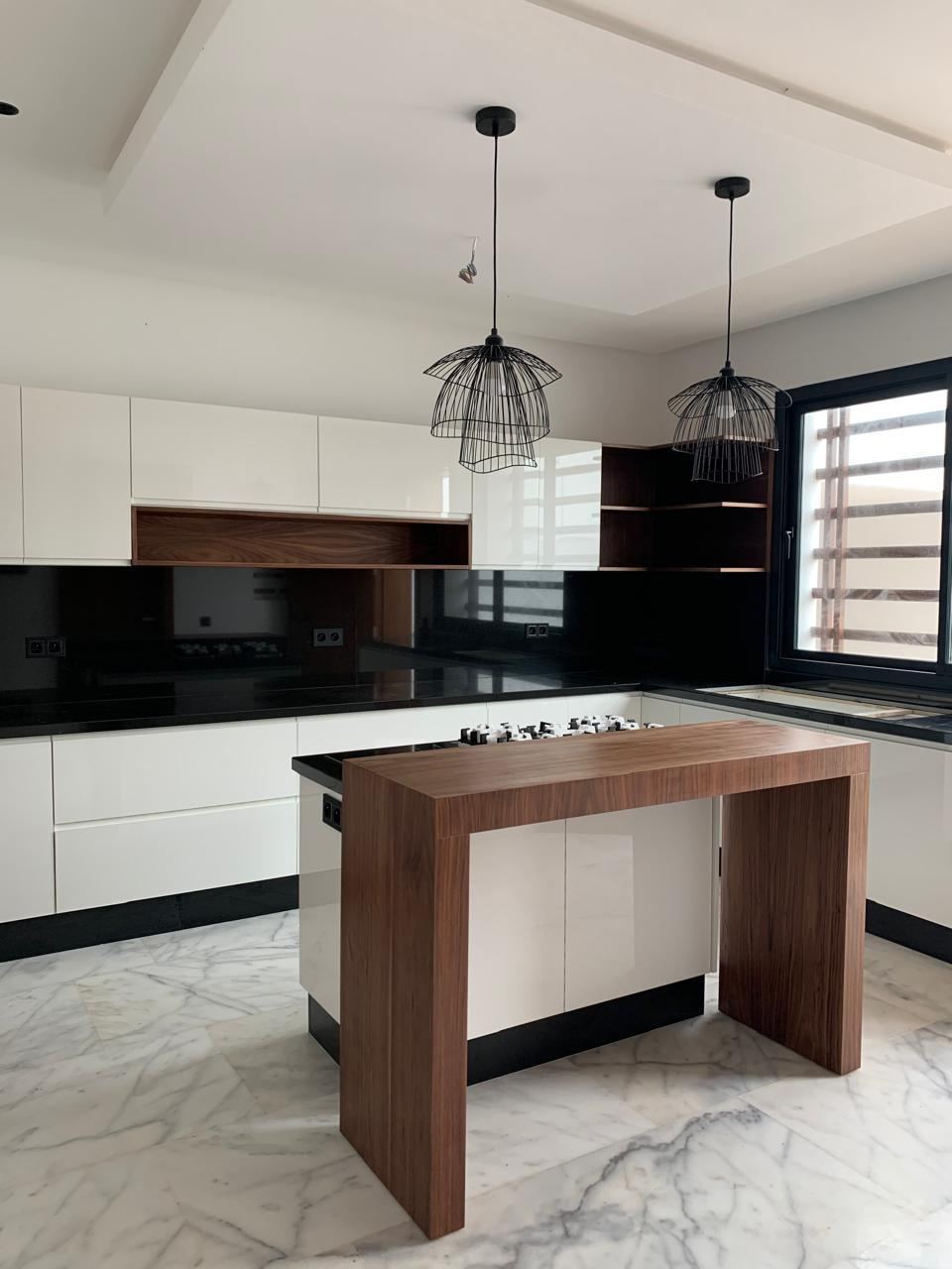 مطبخ على المقاس من إنجاز H&L Menuiserie بالقنيطرة: واجهات بيضاء، وجزيرة مركزية من خشب داكن، وجدار خلفي من الزجاج الأسود، وسطح عمل من الغرانيت.