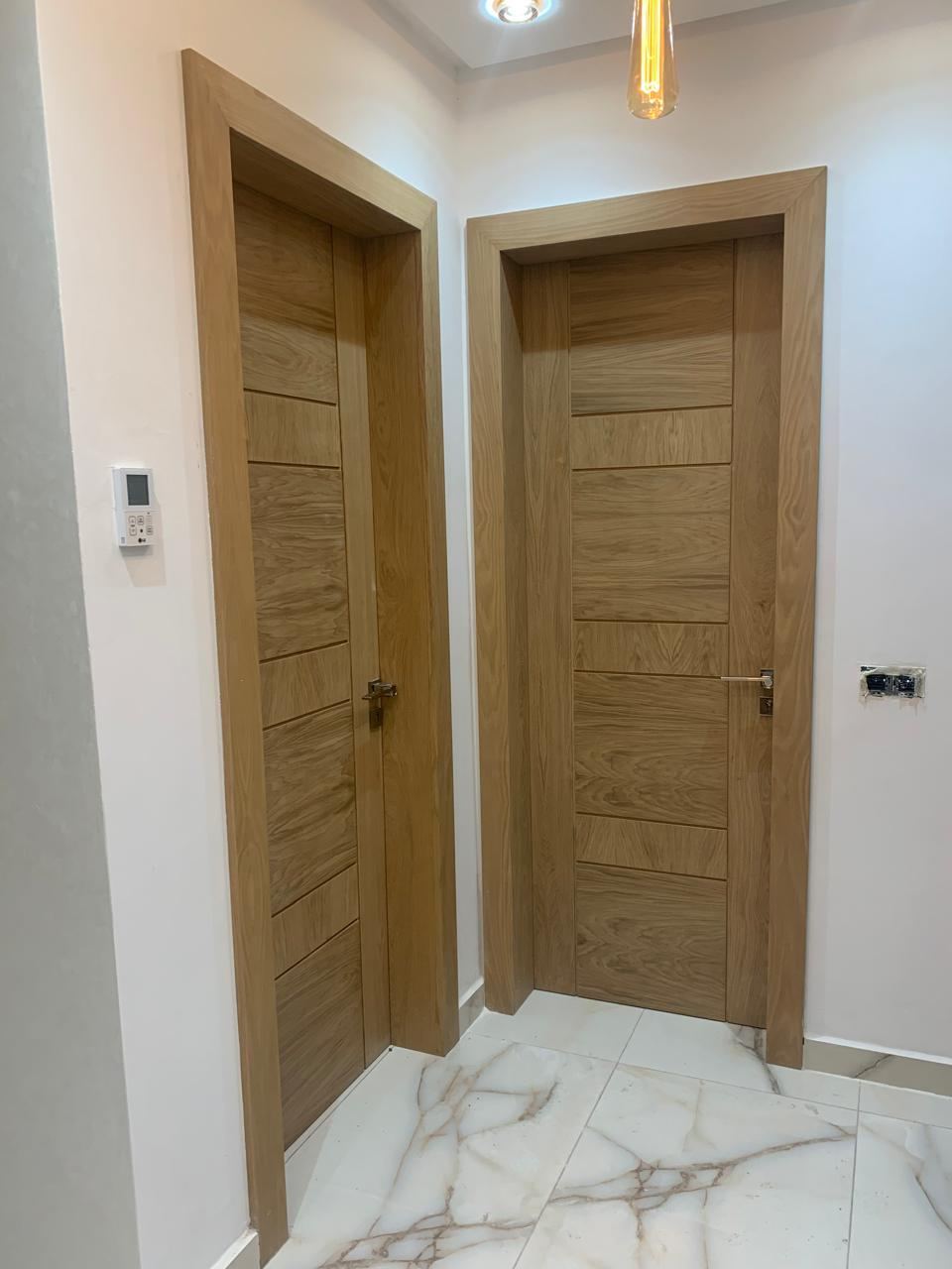 بابان داخليان من خشب فاتح بأخاديد أفقية، مع إطارين متناسقين، على أرضية من الرخام الأبيض المعرّق.